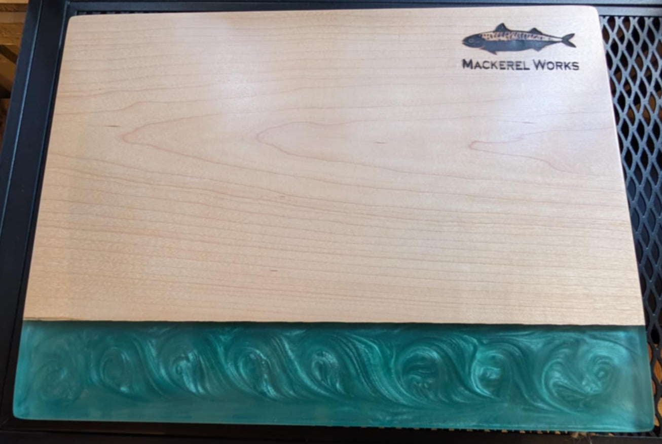 Wooden cutting board with fish design and 'Mackerel Works' branding inside a glass display case.